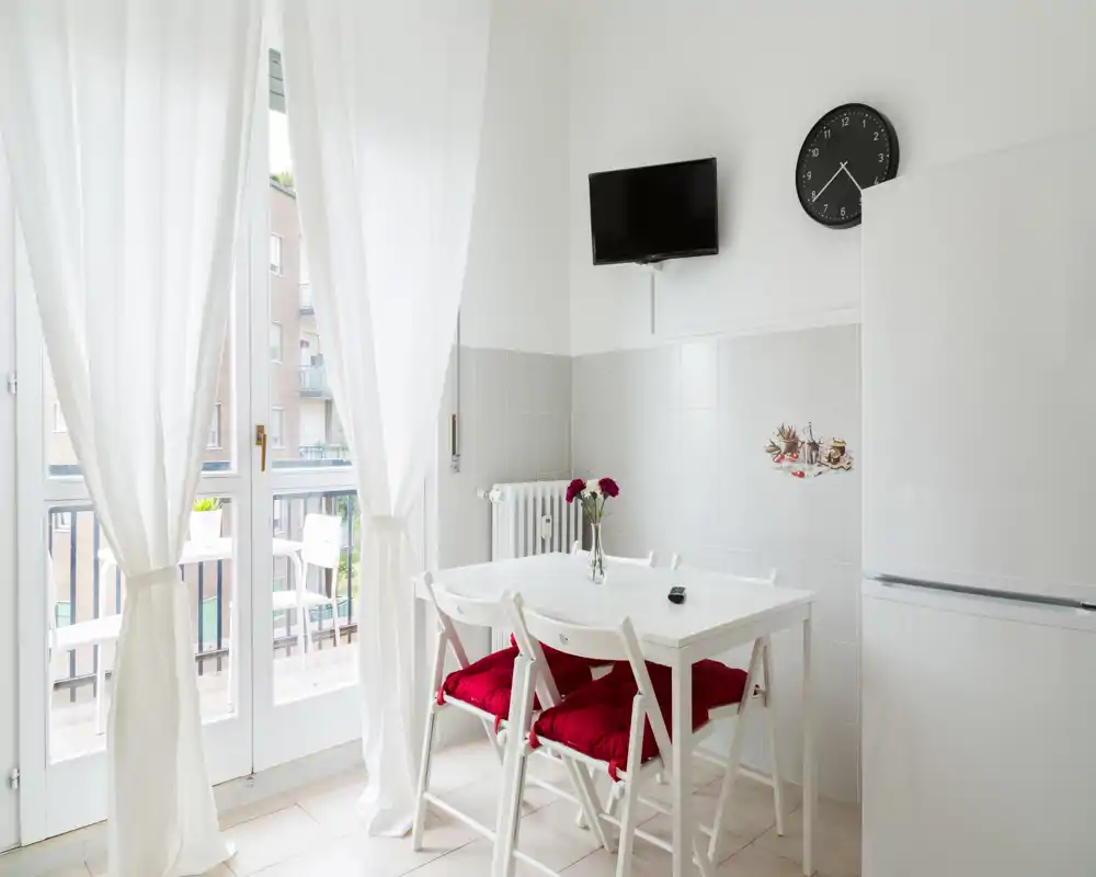 Bright, simple dining area with a small white table, four chairs with red cushions, fresh flowers and large glass doors opening to a balcony.