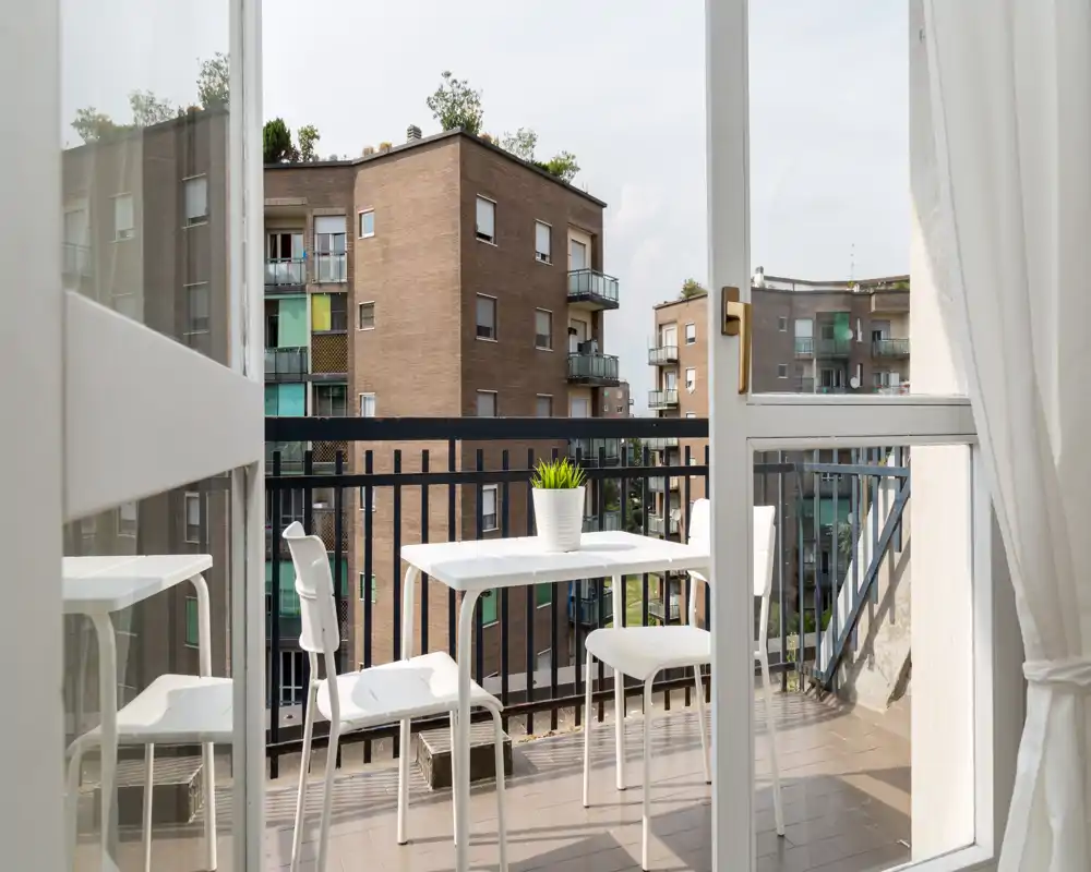 Small city balcony with a white table and chairs, a potted plant, and views of neighboring apartment buildings — bright and inviting for morning coffee.