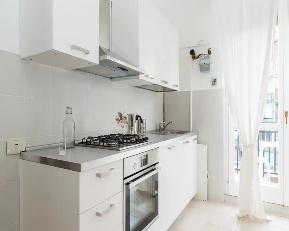 Modern, compact kitchen with white cabinets, built-in oven and gas hob, a stainless countertop and bright natural light from a balcony door.