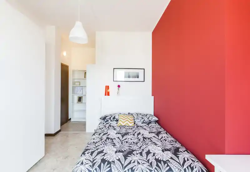 Bright, modern bedroom with a single bed against a bold red accent wall; patterned bedding, bedside lamp and small shelf create a clean, inviting space.