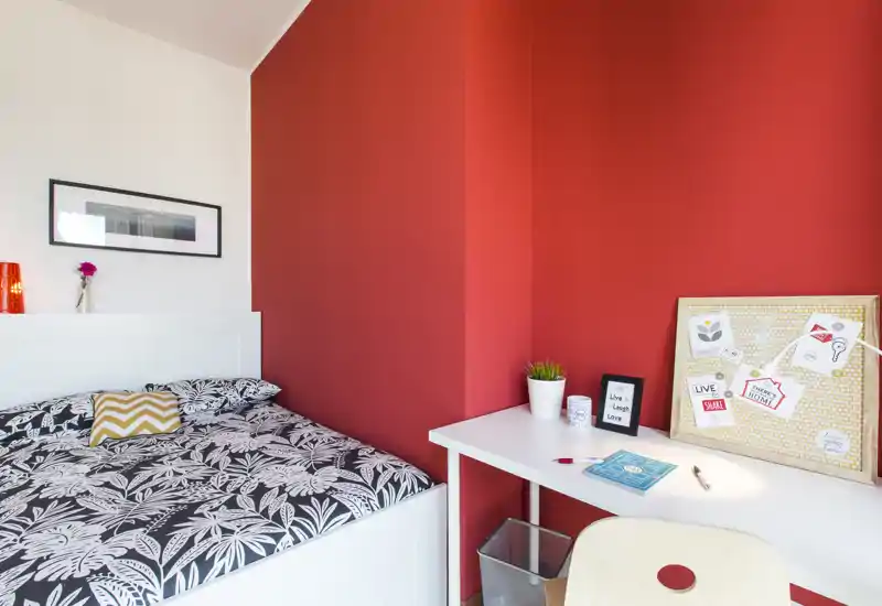 Bright, stylish bedroom with a patterned double bed, white headboard and a tidy desk area against a bold red accent wall — ideal for a modern rental listing.