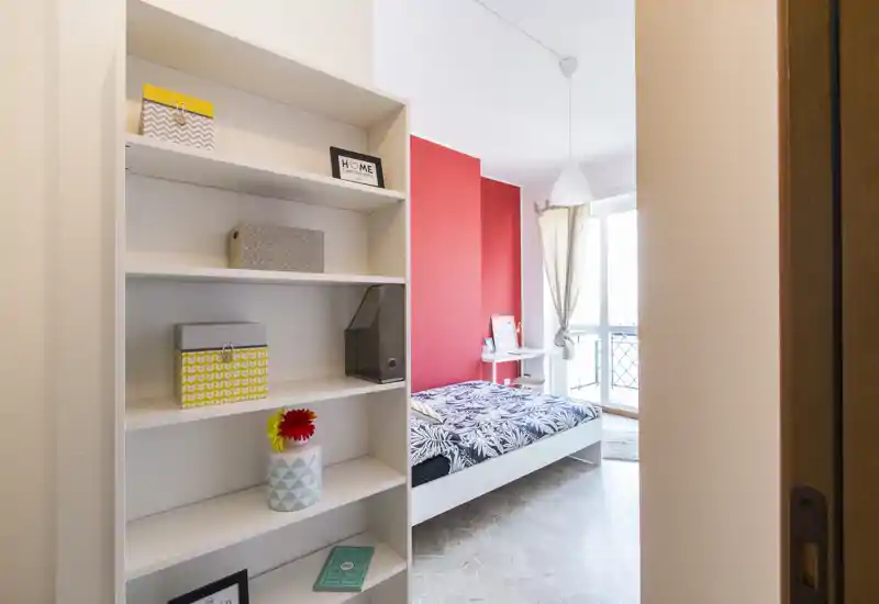 Bright, neatly staged bedroom with a single bed against a red accent wall, natural light from a balcony window and organized shelving in the foreground.