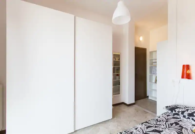Bright, modern bedroom corner with a large white wardrobe and a partially visible patterned bed. The space is clean and simply styled with a pendant lamp and shelving near the doorway.
