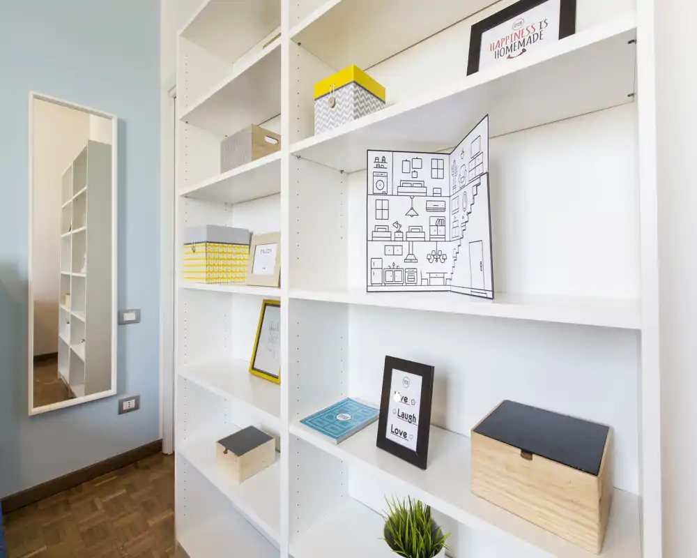 Bright, organized entrance or hallway area with built-in white shelving, decorative boxes and framed accents; a wall mirror reflects the shelf and adds depth.