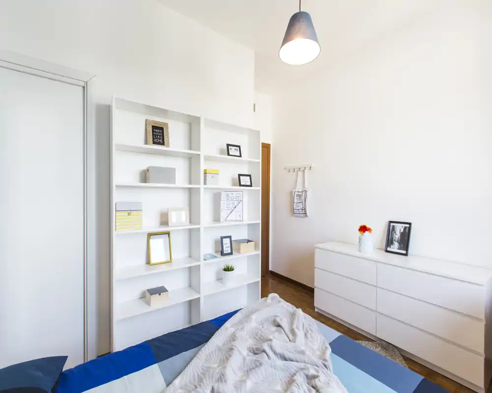 Bright, minimalist bedroom with a neatly made bed, white shelving and a low dresser; clean Scandinavian styling and warm wooden floor.