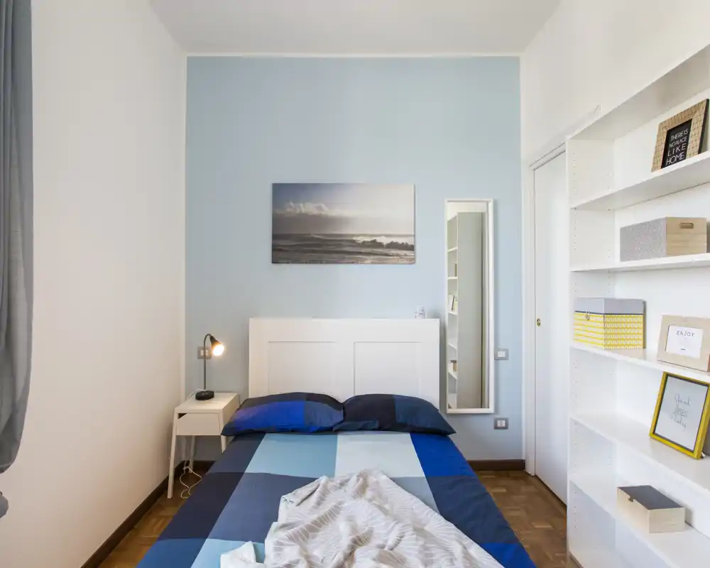 Bright, modern bedroom with a double bed dressed in blue linens, a bedside table with lamp, wall art and a tall mirror; tidy, well-lit and simply styled for rentals.