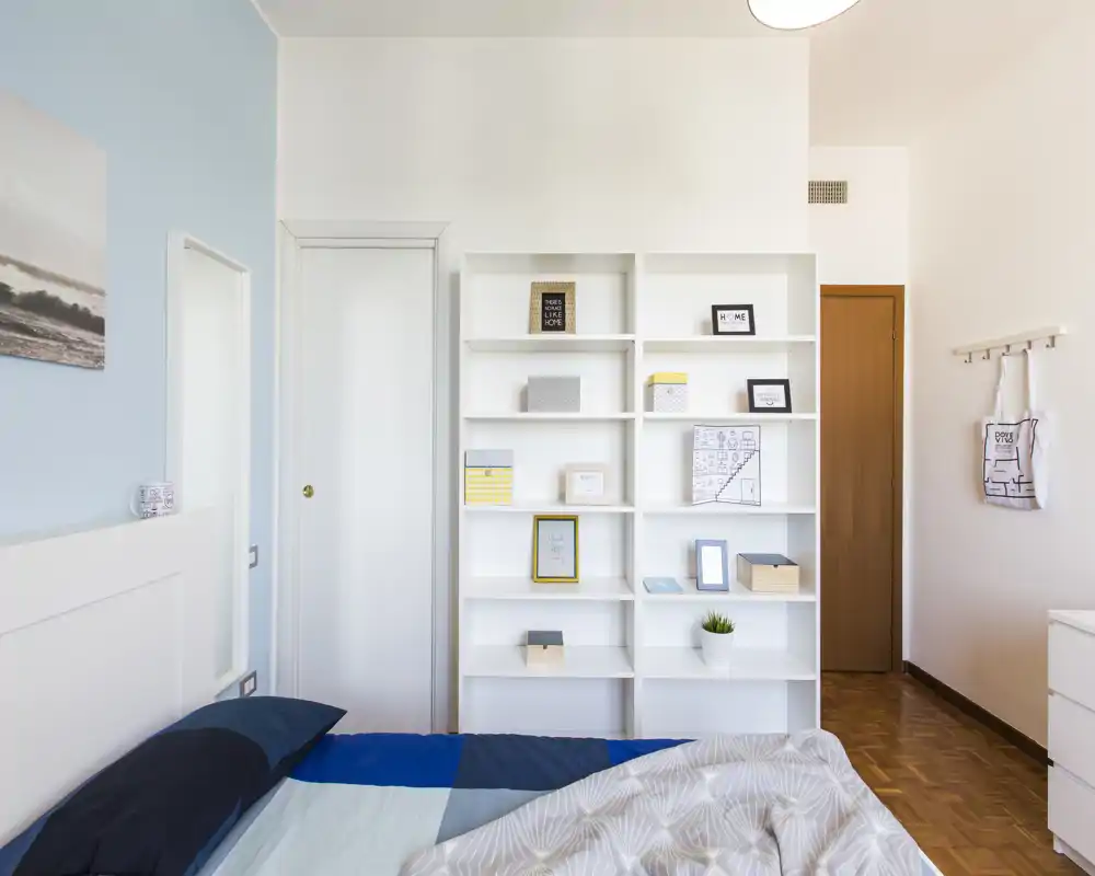 Bright, modern bedroom with a neatly made bed in the foreground and a white shelving unit against the wall; soft blue accent wall and minimalist decor create a calm, airy feel.