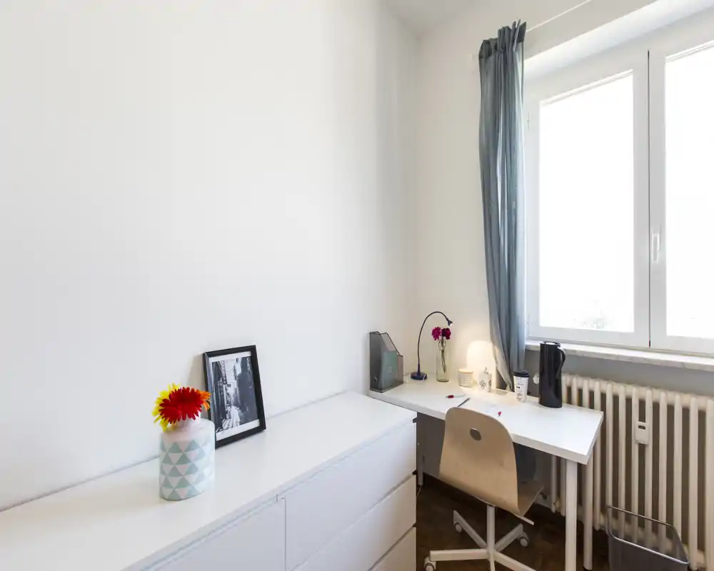 Small, bright home workspace with a white desk by a large window, wooden chair, lamp and minimal decorative touches.