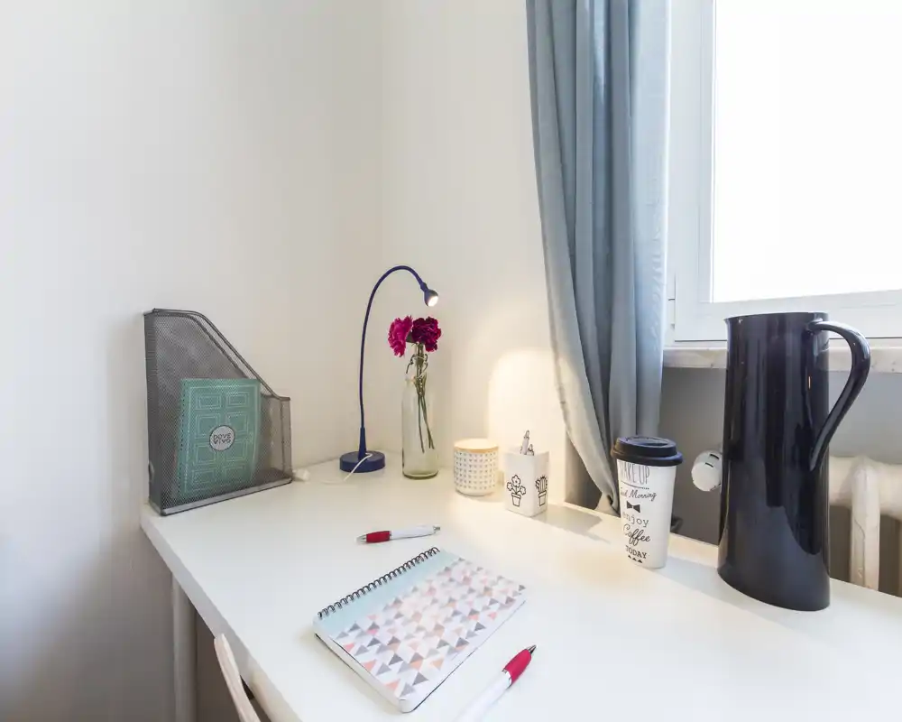 Bright workspace by a window with a white desk, table lamp, notebook and decorative flowers — ideal for a simple study or bedside surface.