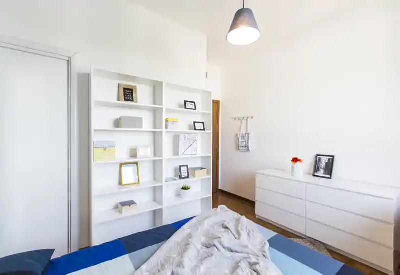 Bright, minimalist bedroom with a neatly made bed, white shelving and a low dresser; clean Scandinavian styling and warm wooden floor.