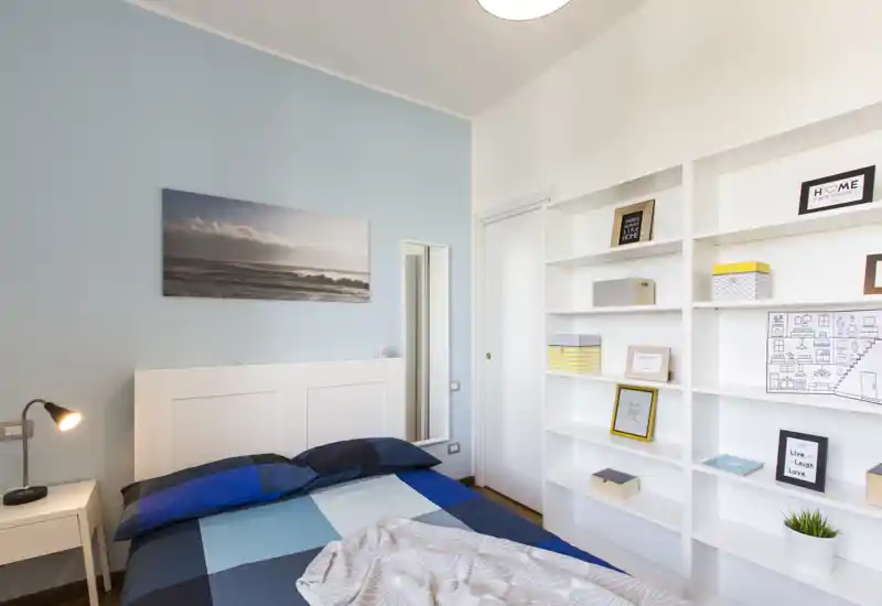 Bright, modern bedroom with a neatly made double bed, bedside table with lamp, a large white shelving unit and soft blue accent wall creating a calm atmosphere.