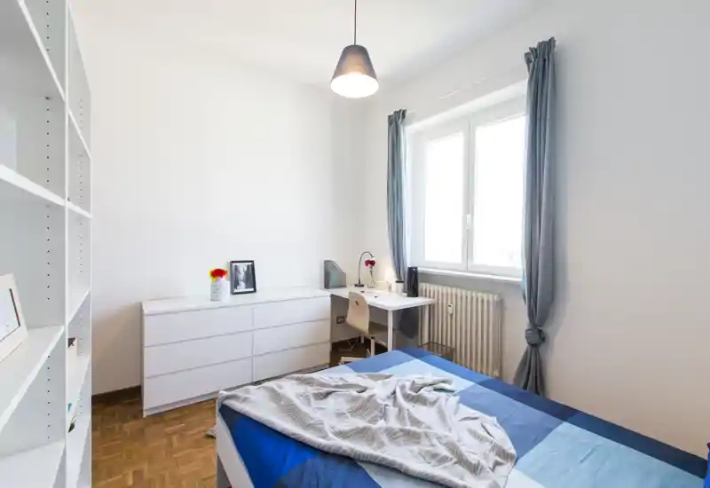 Bright, minimal bedroom with a double bed covered in blue bedding, a white dresser and a small desk by the window — ideal for single or couple renters seeking a tidy, well-lit room.