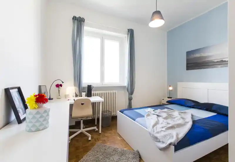 Bright, well-staged bedroom with a double bed, bedside lamp and a tidy desk by the window — clean modern decor and soft blue accents.