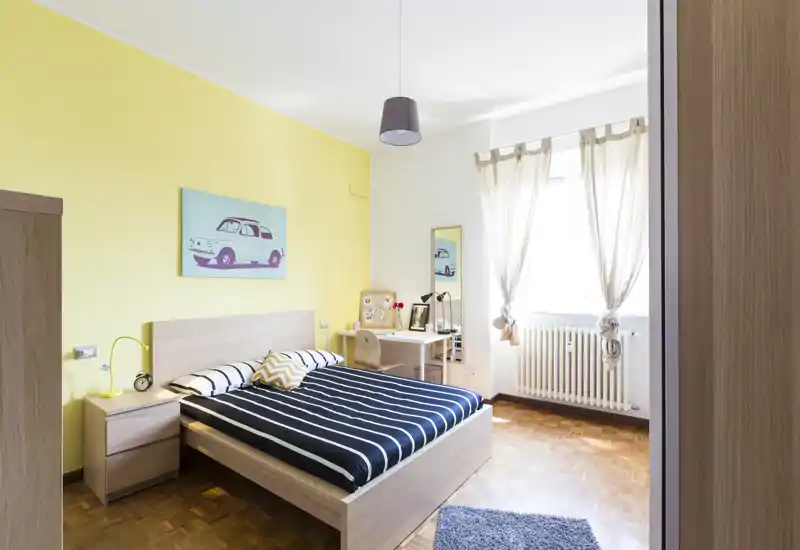 Bright, well-styled double bedroom with a wooden bed, bedside table and a small desk by the window. The room features fresh yellow accent wall, parquet flooring and natural light, ideal for a cozy rental listing.