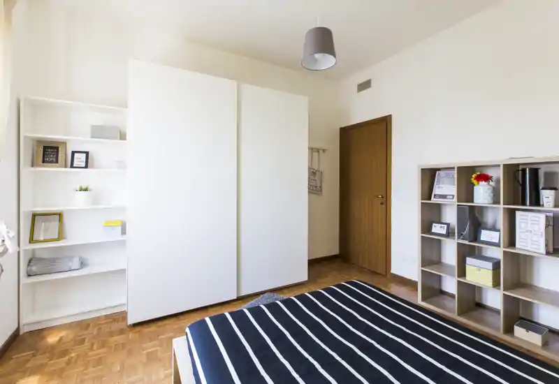 Bright, modern bedroom with a striped bed in the foreground, white wardrobes and open shelving providing storage and decorative accents.