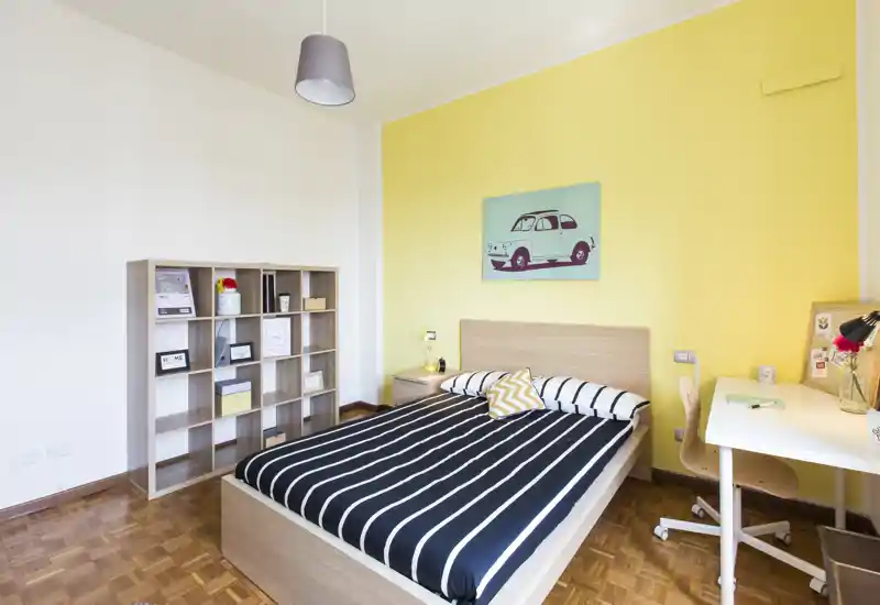 Bright, well-staged bedroom with a double bed, striped bedding, a nightstand and a desk; cheerful yellow accent wall and neat shelving create a welcoming, modern feel.
