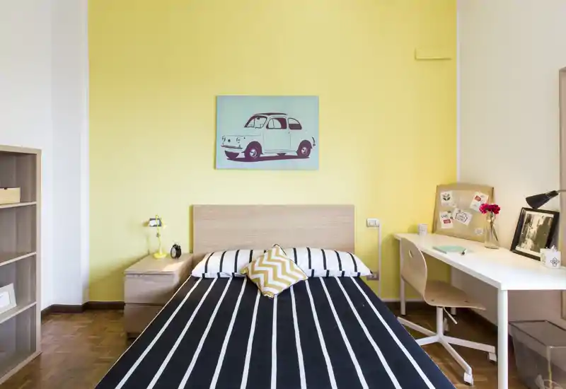 Bright, modern bedroom with a double bed centered against a cheerful yellow accent wall, bedside table and a functional desk — ideal for a student or professional.