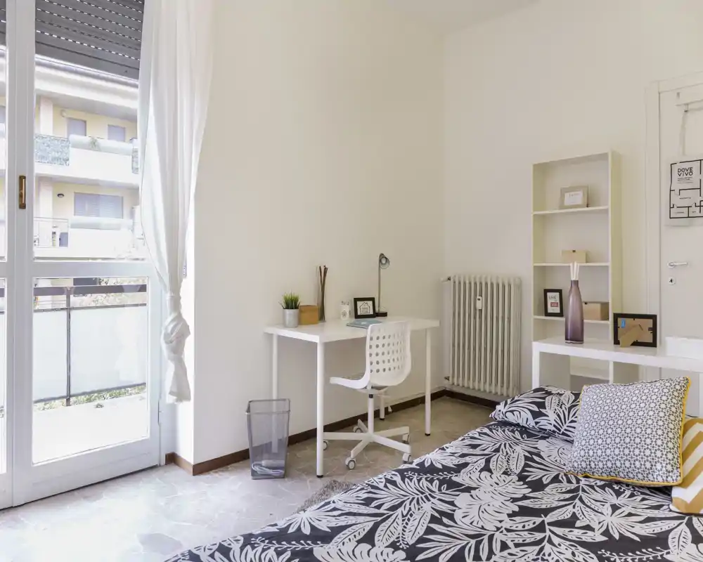 Dormitorio luminoso y minimalista con colcha estampada, cojines decorativos, escritorio blanco y silla junto a la puerta del balcón que aporta mucha luz natural — ideal para descansar y trabajar.