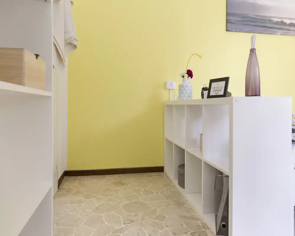 Pequeño rincón de salón ordenado con estantería blanca modular, jarrón decorativo y marco en pared amarillo claro; luz natural y suelo de piedra que aportan un ambiente fresco y minimalista.