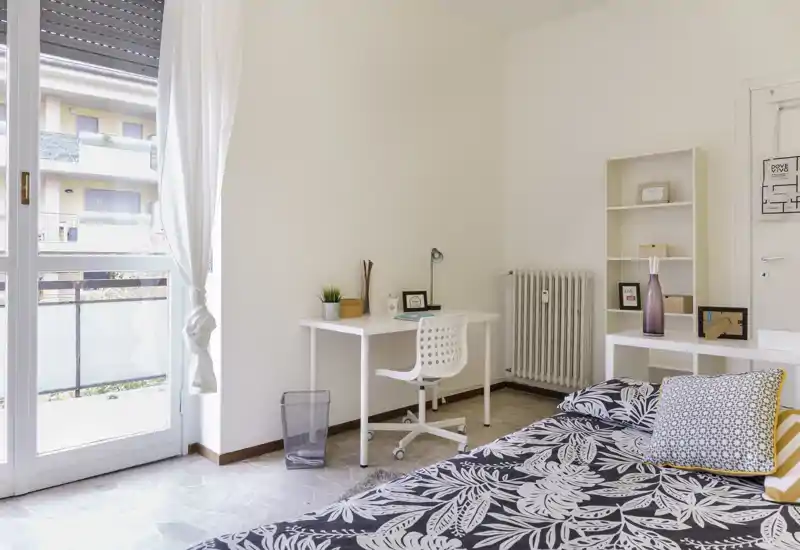 Dormitorio luminoso y minimalista con colcha estampada, cojines decorativos, escritorio blanco y silla junto a la puerta del balcón que aporta mucha luz natural — ideal para descansar y trabajar.