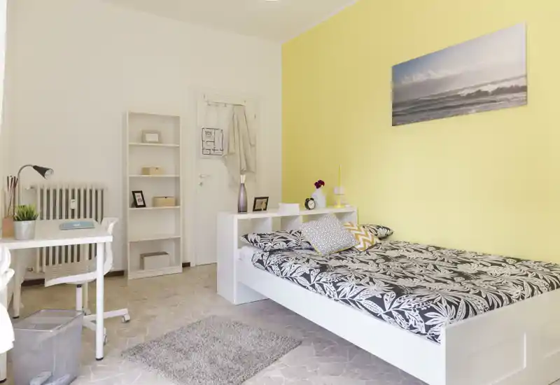 Dormitorio luminoso y ordenado con cama baja blanca, ropa de cama estampada y pared amarilla. La habitación cuenta con escritorio, estantería y una decoración cuidada, ideal para un estudiante o una persona sola.