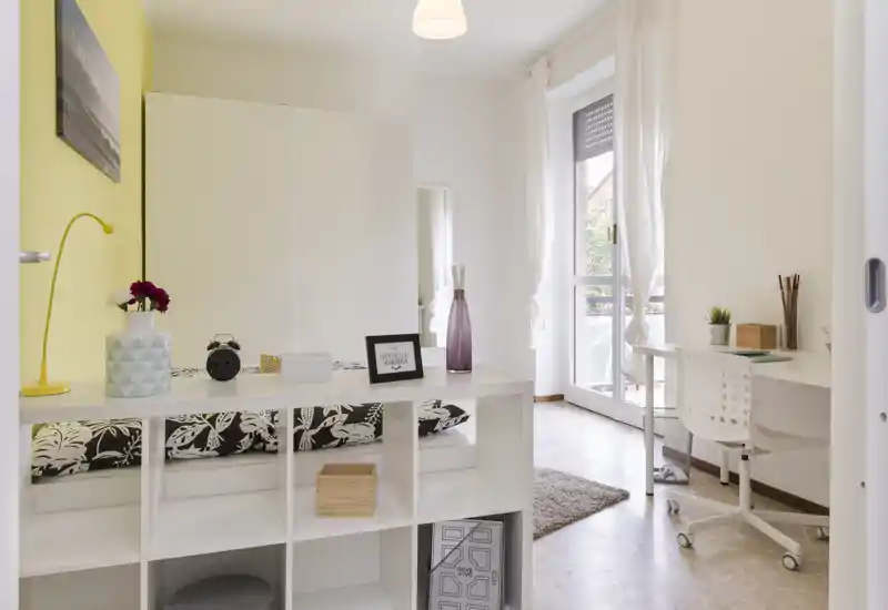 Dormitorio moderno y luminoso con estantería a los pies de la cama, cojines estampados, escritorio junto a la puerta del balcón y agradable luz natural — bien preparado para alquileres.