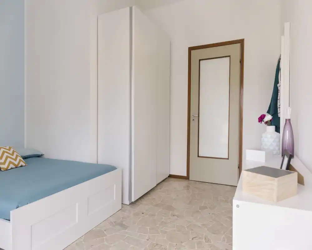 Camera da letto luminosa e pulita con struttura del letto bianca e biancheria turchese. Armadio alto bianco, pavimento in piastrelle e arredi minimal per un'atmosfera tranquilla.