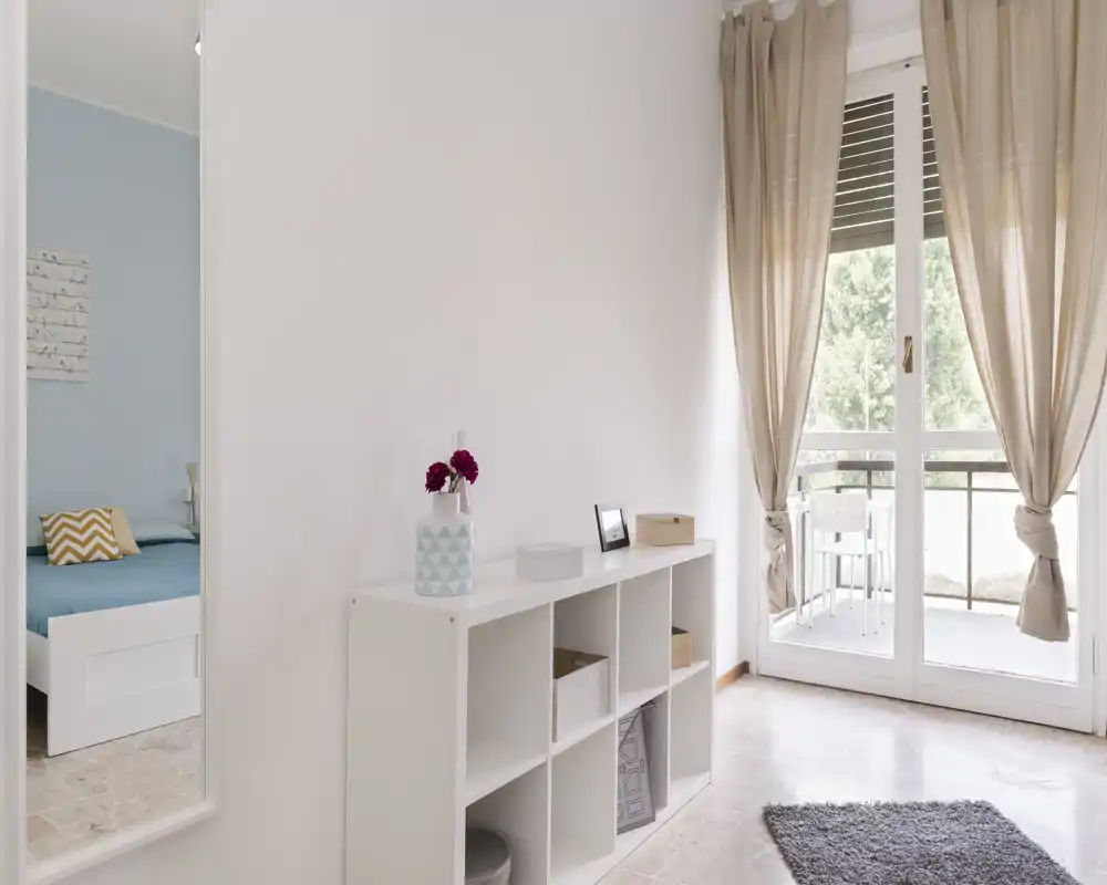 Ampia vista luminosa verso la camera da letto attraverso una porta aperta, con struttura del letto bianca, cuscini decorativi e luce naturale dal balcone. Arredamento neutro e piccolo mobile contenitore rendono l’ambiente pulito e arioso.