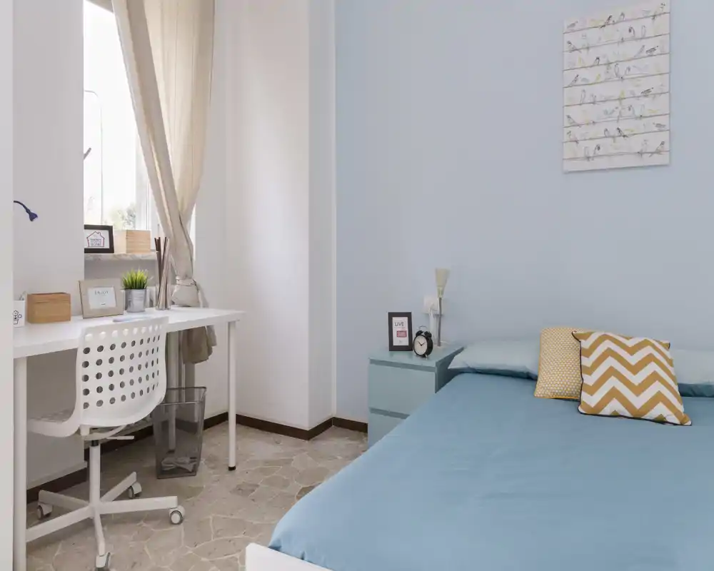 Camera da letto luminosa e accogliente con letto matrimoniale ben rifatto in lenzuola blu, comodino con lampada e cuscini decorativi; piccolo angolo studio vicino alla finestra con luce naturale.