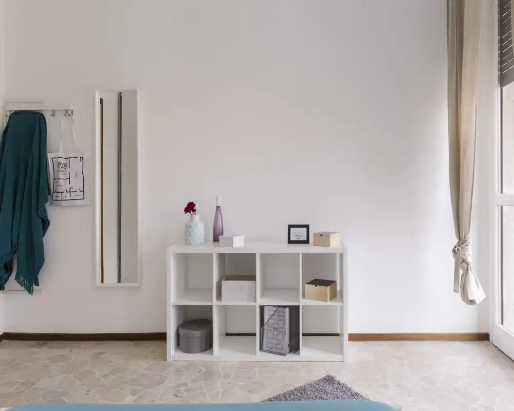 Angolo di camera minimale con un mobile contenitore a cubi bianco, oggetti decorativi e luce naturale da una grande finestra; ordinato, luminoso e dallo stile semplice.