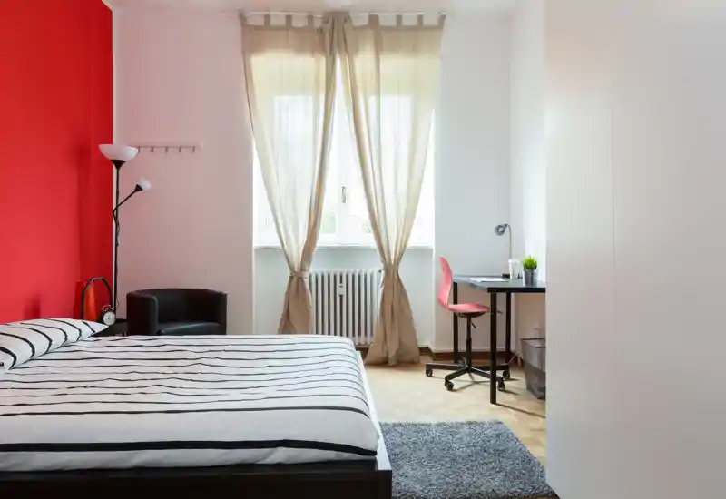 Dormitorio moderno y luminoso con cama doble a rayas, pared de color rojo como acento y un pequeño escritorio junto a la ventana — ideal para una o dos personas.