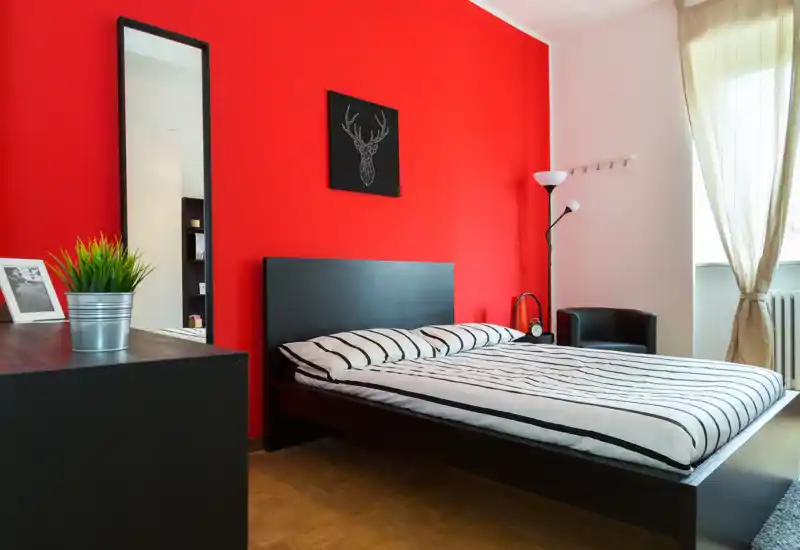Dormitorio moderno y luminoso con cama de madera negra frente a una pared roja intensa, ropa de cama a rayas y luz natural desde la ventana. Mobiliario minimalista y con estilo: espejo alto, lámpara de pie y sillón pequeño.