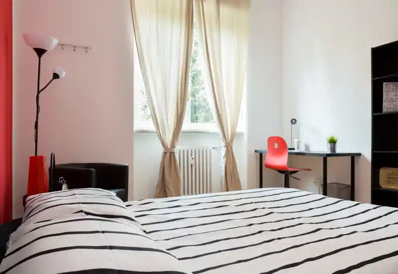 Dormitorio moderno y luminoso con cama grande a rayas, luz natural desde la ventana central y una pequeña zona de trabajo con silla roja — acogedor y con estilo.