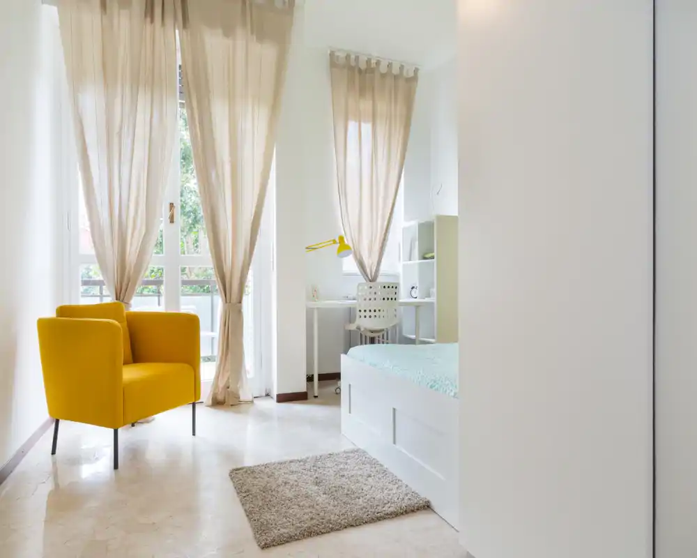 Bright, airy bedroom with a comfortable white bed, a cozy beige rug and a vibrant yellow armchair by large windows that let in abundant natural light.