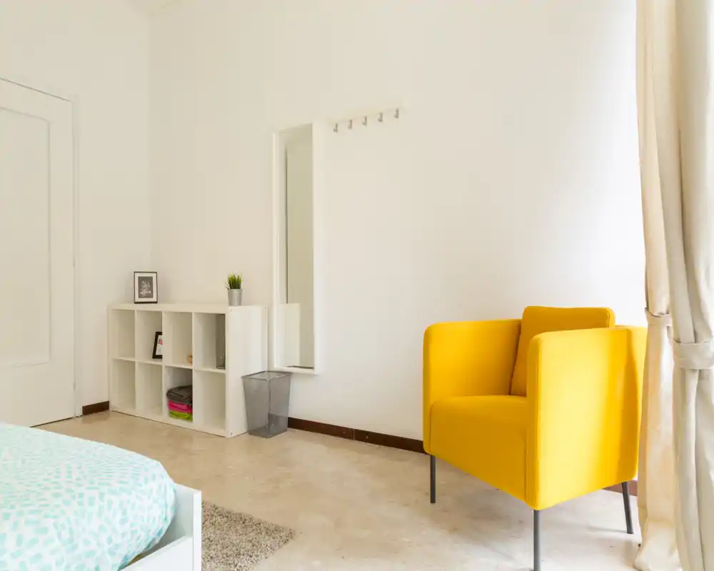 Bright, minimalist bedroom corner with a patterned bed edge, open shelving, a full-length mirror and a vivid yellow armchair providing a warm focal point.