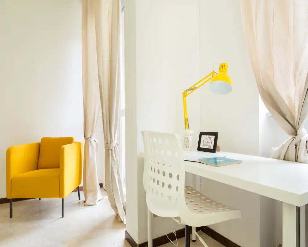 Bright, modern living area corner with a yellow accent armchair, white desk and chair near a window, and a matching yellow desk lamp — ideal for relaxing or working by natural light.
