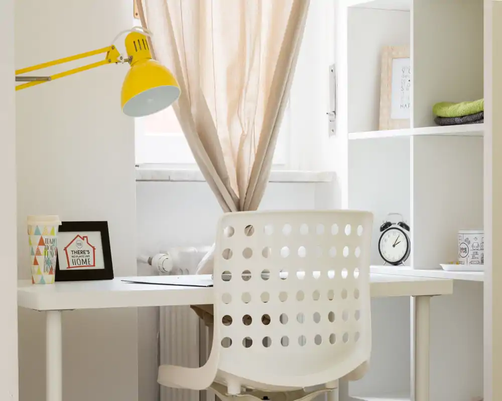 Bright, minimal home workspace with a white desk, perforated chair, yellow desk lamp and shelving; tidy and naturally lit, suitable for focused work or study.