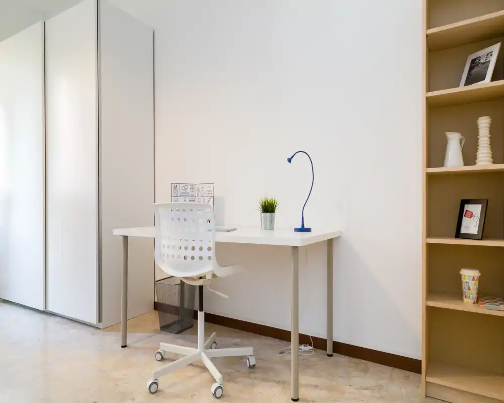 Zona de estar luminosa y minimalista con un escritorio sencillo: mesa y silla blancas, lámpara de escritorio azul y estantería para almacenamiento—ideal como rincón de oficina en casa.