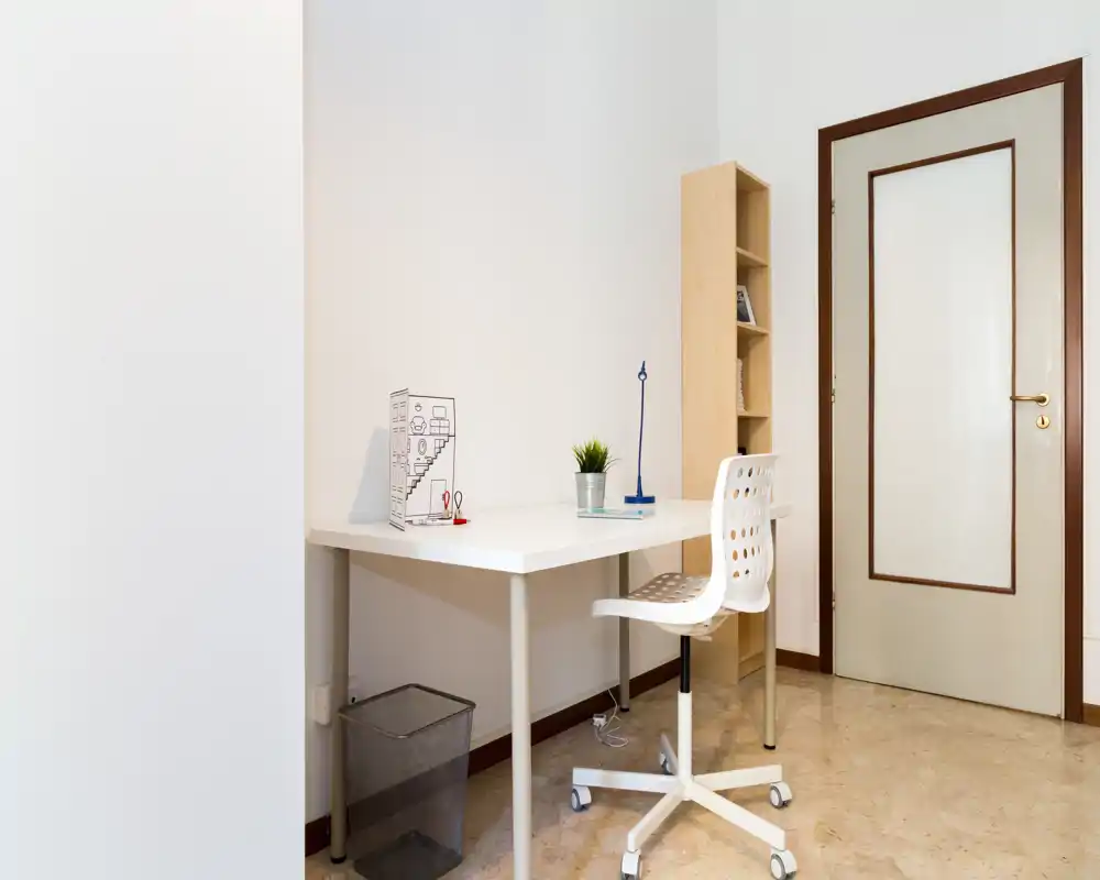 Rincón de oficina en casa minimalista con escritorio blanco, silla giratoria, pequeño estante y planta; espacio de trabajo luminoso y ordenado.