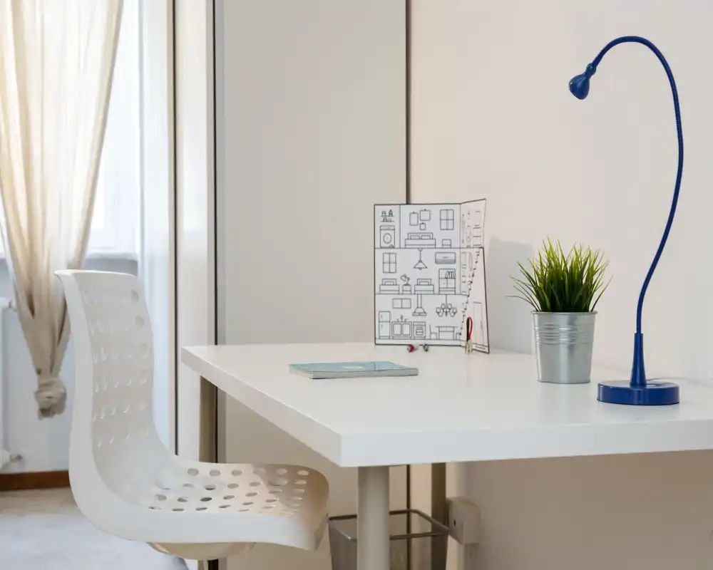 Rincón de estudio luminoso y minimalista con escritorio blanco, silla moderna perforada, lámpara azul y una pequeña planta junto a la ventana.