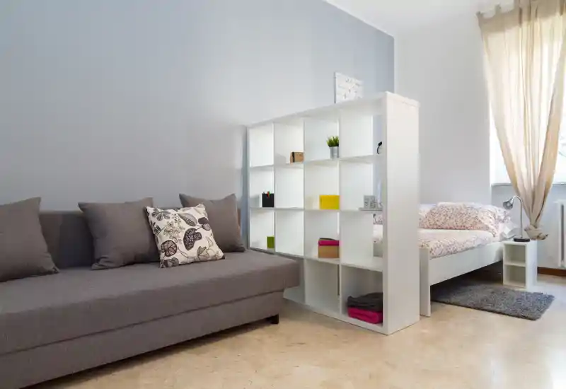 Rincón de dormitorio luminoso y ordenado con una cama individual detrás de un estante abierto y un sofá gris en primer plano. La luz natural de la ventana brinda una atmósfera acogedora.