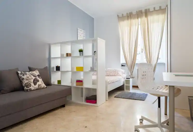 Dormitorio moderno y luminoso con cama individual junto a una gran ventana, una estantería blanca de cubos como separador, un sofá gris y un pequeño escritorio. La luz natural y los tonos neutros crean un ambiente tranquilo y aireado, perfecto para un anuncio de alquiler.