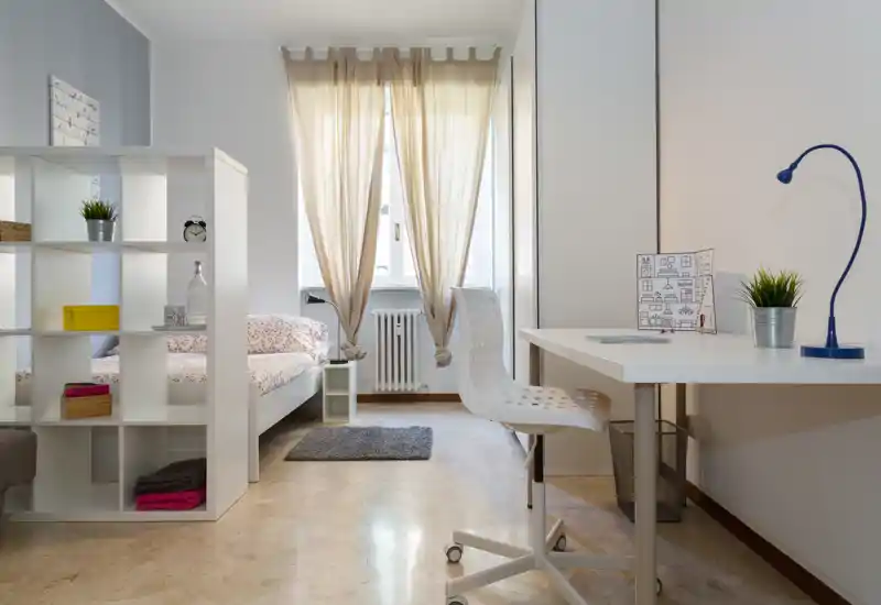 Dormitorio moderno y luminoso con cama individual parcialmente visible detrás de una estantería blanca, escritorio ordenado con silla y lámpara, y suave luz natural desde la ventana.