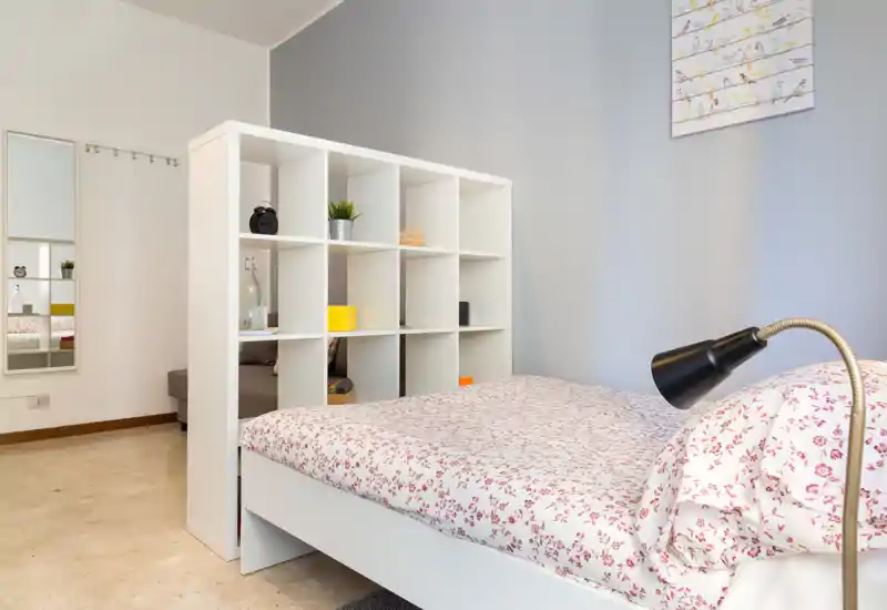 Dormitorio moderno y luminoso con edredón de flores y una estantería blanca que separa la cama del área de estar; líneas limpias y tonos suaves que transmiten tranquilidad.