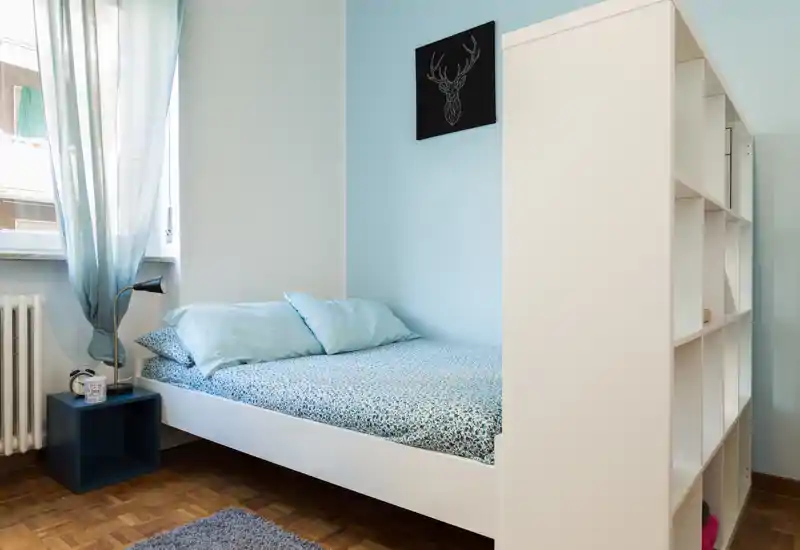 Bright, neatly staged single bedroom with a white bedframe, patterned linen and soft blue accents. Natural light from the window highlights wooden parquet flooring and simple shelving, creating a calm, welcoming space ideal for listings.