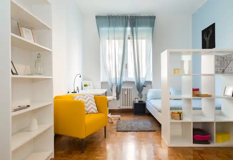 Bright, modern bedroom-studio with a cozy bed tucked behind an open shelving divider, a cheerful yellow armchair, and a small desk by the window letting in abundant natural light.