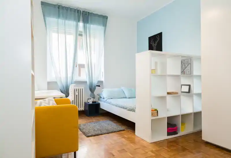 Bright, cozy single bedroom featuring a low white bed with soft blue bedding, a decorative shelving divider and a sunny window framed by sheer curtains.