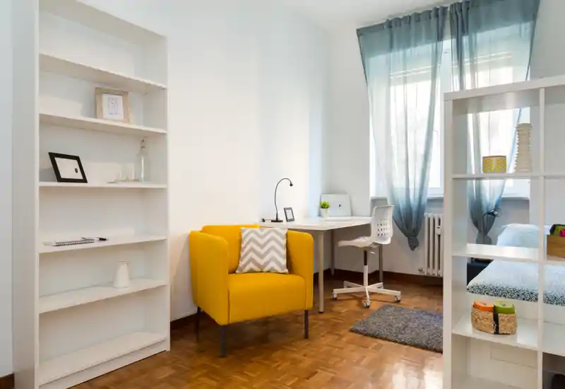 Bright, modern bedroom corner with a bold yellow armchair, a desk by the window and a partially visible bed separated by shelving; clean parquet floor and soft natural light create a welcoming atmosphere.
