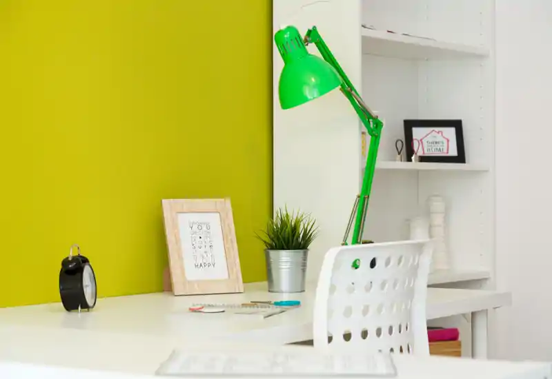 Bright, modern study corner with a white desk, perforated chair and a green adjustable desk lamp against a lime accent wall. Decorative touches like a small plant and framed print create a fresh, inviting workspace.