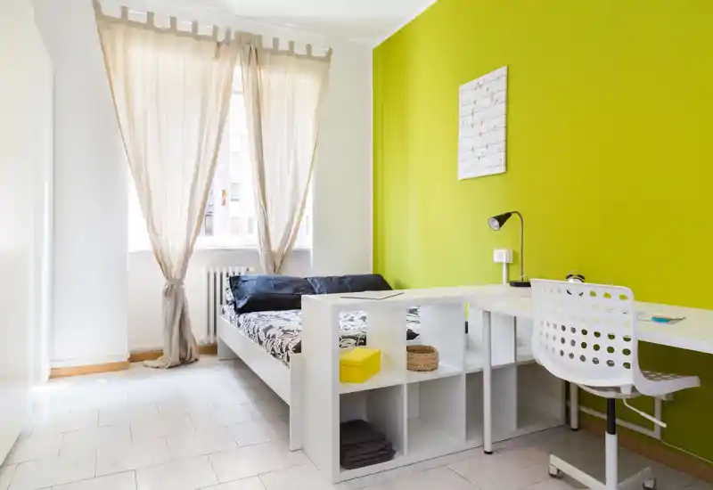 Bright, modern bedroom with a double bed, natural light from a tall window and a white desk with shelving — ideal for a student or single renter.